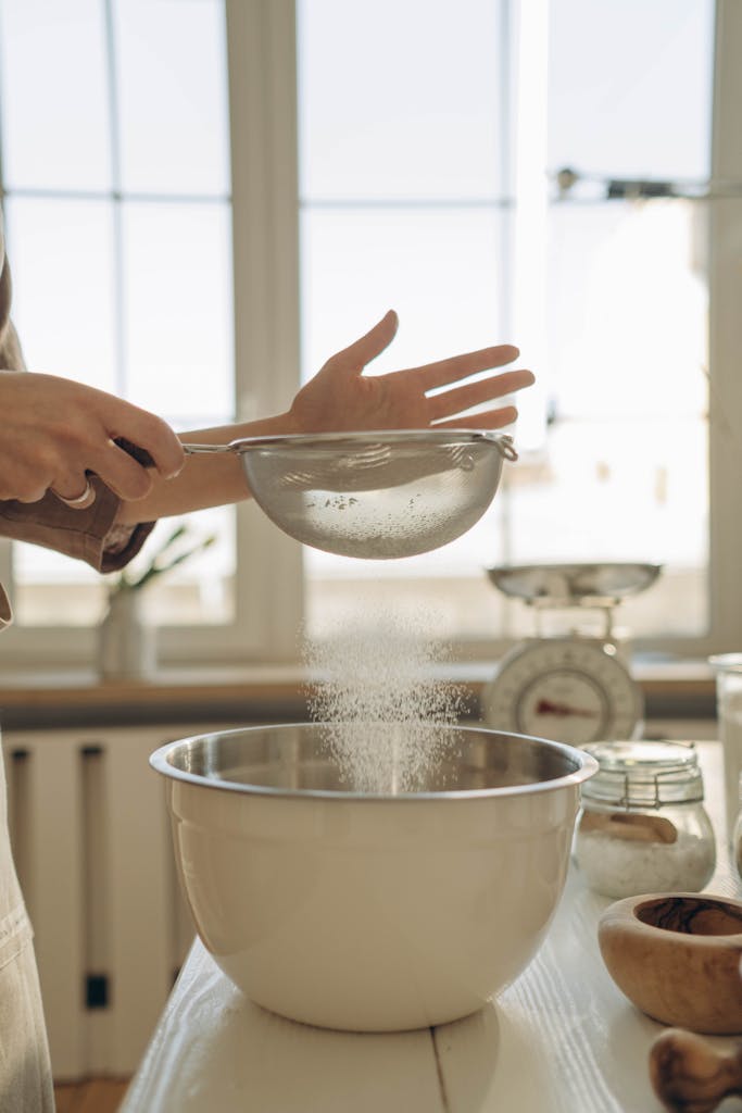 Close-up of hands sifting flour into a mixing bowl for baking in a sunny kitchen.