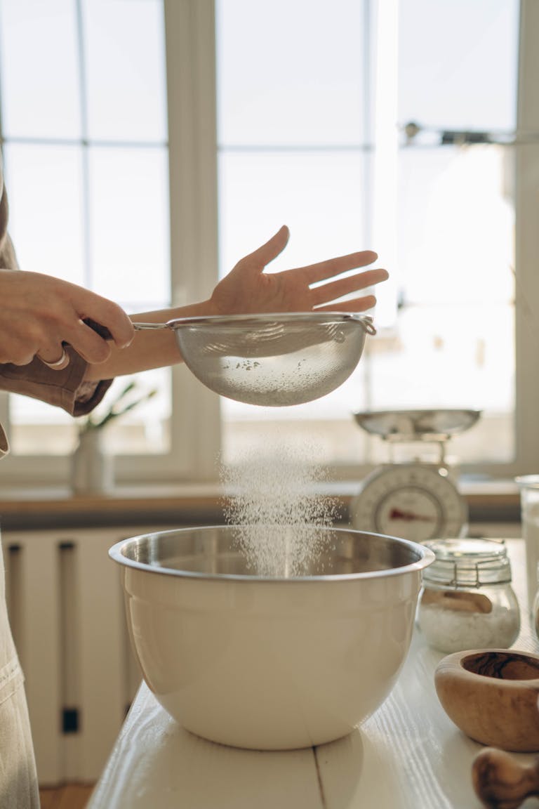 Close-up of hands sifting flour into a mixing bowl for baking in a sunny kitchen.