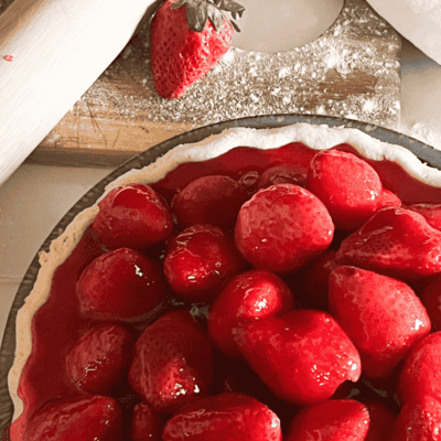 Freshly made glazed strawberry pie sitting on a table next to a cutting board with a rolling pin.