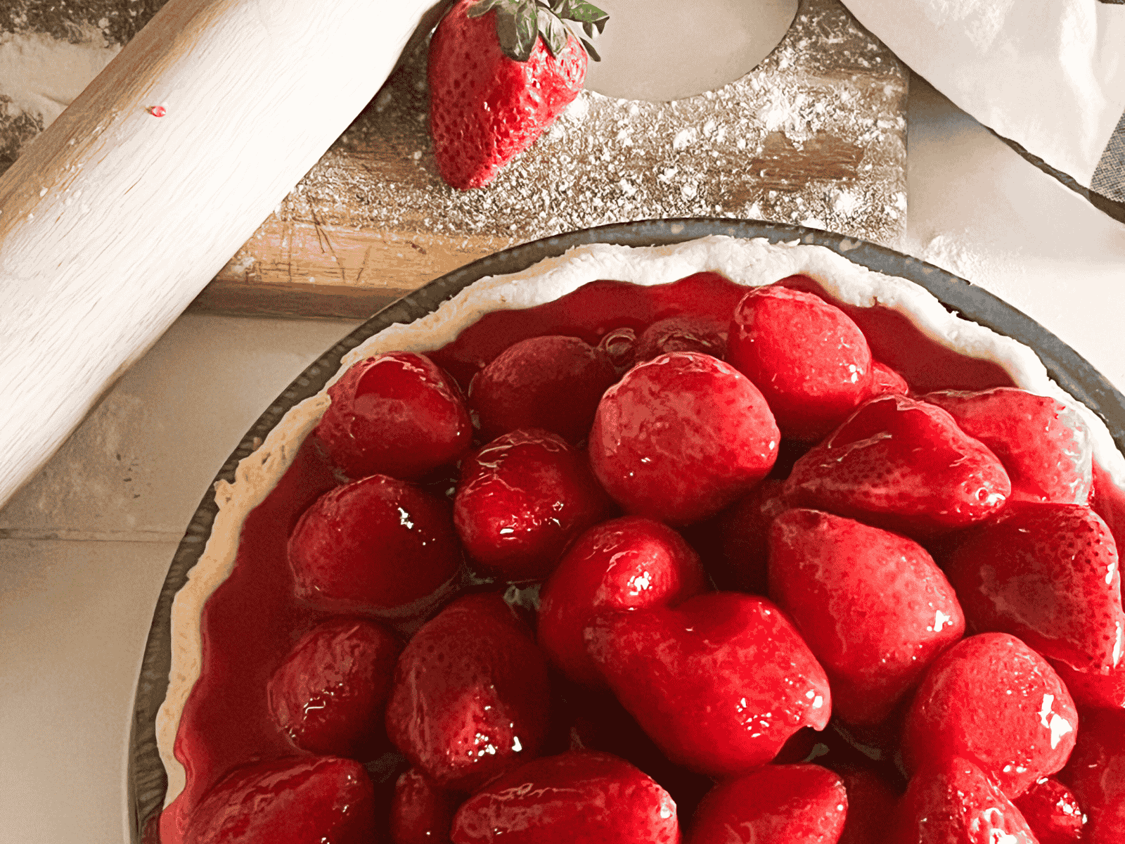 Freshly made glazed strawberry pie sitting on a table next to a cutting board with a rolling pin.