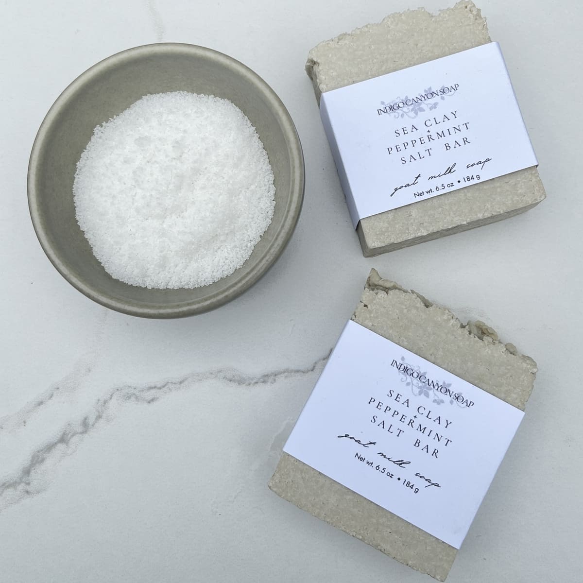 Two gray salt bars sitting on a white marble table with a gray bowl of white sea salt sitting next to them.