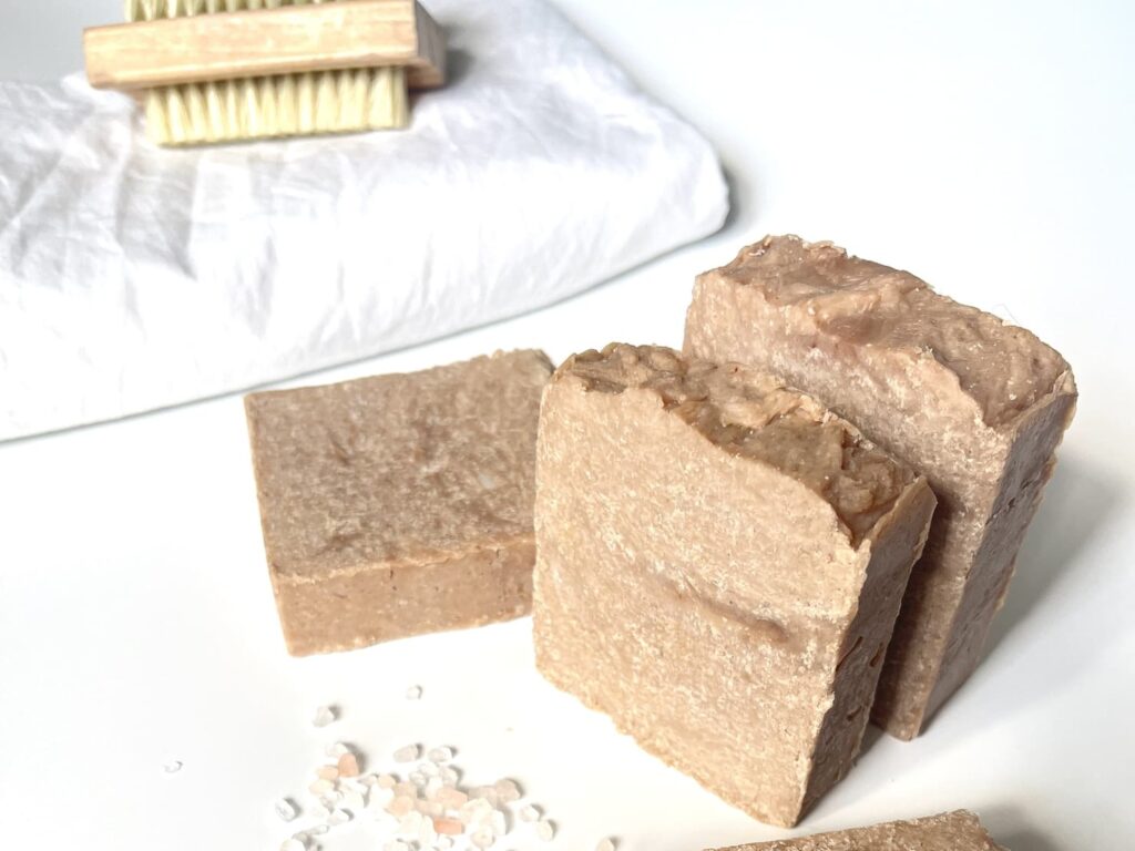Bars of hot process salt soaps sitting on a white table with pink salt around it and towels and a natural bristle brush in the background.